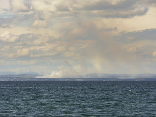 Smoke from a fire, view from a sea. Cloudy day, blue water, clouds.