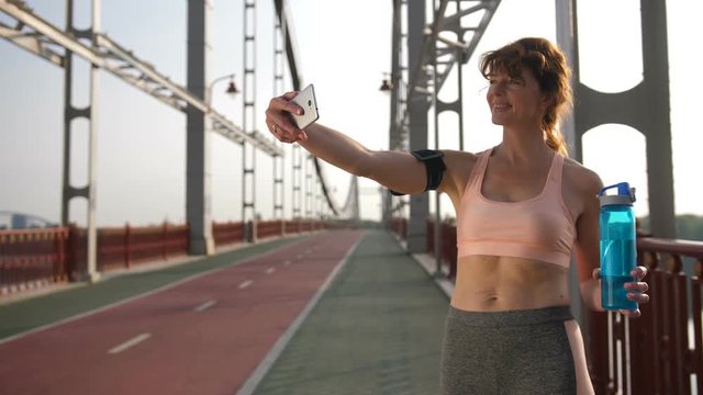 Fitness selfie senior redhead woman taking self portrait photograph with smart phone after running exercise workout on city bridge during sunrise. Healthy lifestyle with fit and sporty senior woman.