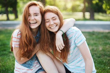 Wanna hug her forever. Portrait of happy and carefree two twin sisters with natural red hair and freckles, laughing out loud and cuddling, fooling around while resting in park on fresh green grass - obrazy, fototapety, plakaty