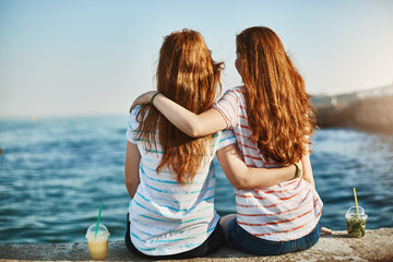 Girs making promises never leave each other, gazing at beautiful sea and hugging, sitting near docks, dreaming about future, talking casually like sisters and enjoying warm sunny evening together