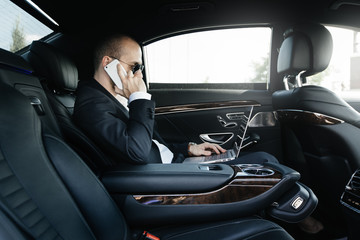 Confident young businessman working on his laptop and talking on the phone while sitting on the back seat of luxury car