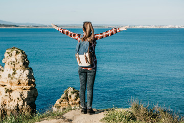 A tourist girl or traveler with a backpack admires the beautiful view of the Atlantic Ocean in Portugal and raises her hands up showing how she likes and how she is free.