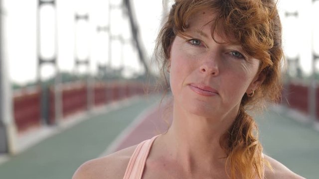 Closeup portrait of beautiful senior redhead sporty woman looking at camera and playing with hair after running exercise workout on the bridge. Wise and peaceful eyes of attractive female in her 40s.