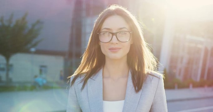 Portrait of the young charming woman in glasses standing outdoor in the sunlight and smiling while turning face to the camera.