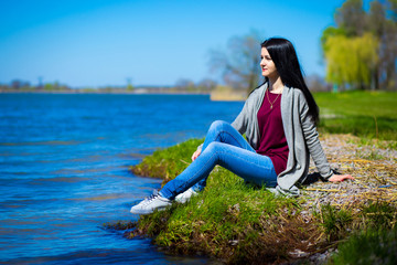 Fototapeta na wymiar Beautiful young woman in jeans sits on river bank in sunny day