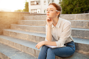 beautiful young redhead girl with freckles sitting on a stairs and making notes in a notebook. Portrait of a student. Remote education. Homework