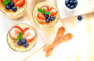 homemade, exquisite dessert tiramisu in glasses decorated with strawberry, blueberry, mint on white wooden table, top view.