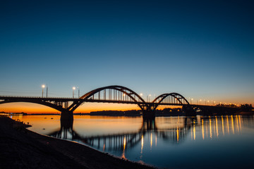 Naklejka premium Volga bridge over Volga river after sunset with reflection in water, Yaroslavl region, Rybinsk city, Russia. Beautiful night landscape