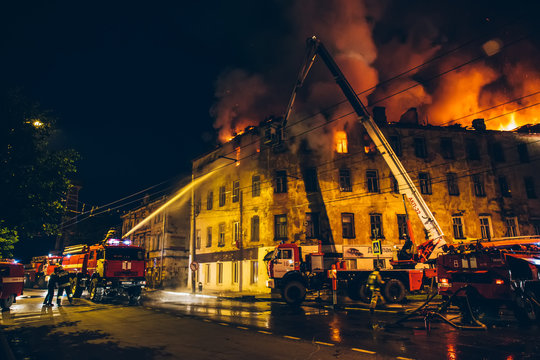 Firefighters With Fire Hose And Fire Engines Or Trucks Are Fighting Fire In Residential Building At Night. Huge Smoke, Flame In Burning House