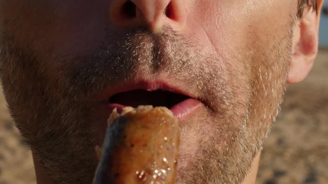 Closeup of unshaven man eating appetizing grilled sausage outdoors