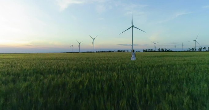 Beautiful girl walking on a green wheat field with windmills for electric power production
