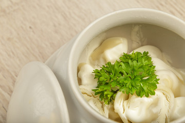 Dumplings in a white pot with sour cream and greens on a wooden background.