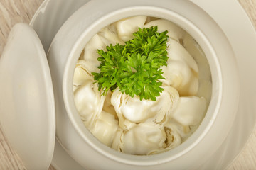 Dumplings in a white pot with sour cream and greens on a wooden background.