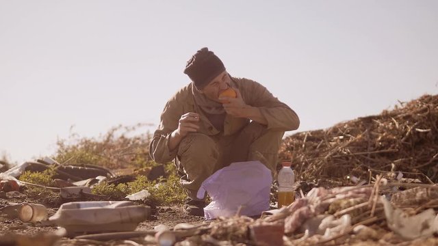 portrait of a dirty homeless hungry man in a dump eating orange for food in the package with walking goes looking for food slow motion video. homeless dirty man roofless person looking for food in a