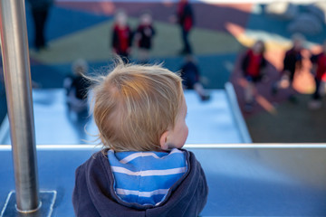 A young child at the top of the metal slide watches other children playing and sliding down
