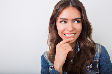 Fototapeta premium Close-up portrait os funny charming girl with long dark hair and lovely smile playful looking at camera and bits lips on the background of white wall
