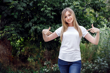 Smiling beautiful woman pointing at her empty white T-shirt with both index fingers, copy space for...