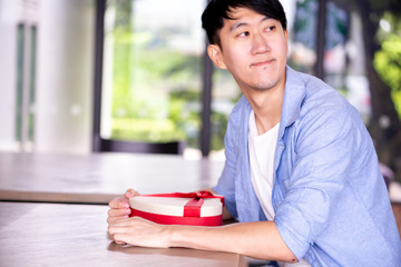 Young Asian man patiently sitting in cafe restaurant and holding a present gift giving to someone...