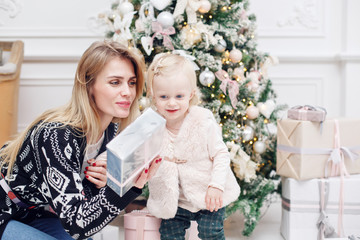 Obraz na płótnie Canvas Cheerful mom and her cute daughter baby open a gift. Parent and little child having fun near Christmas tree indoors. Loving family Merry Christmas and Happy New Year.