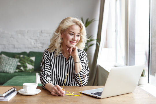 Elderly Woman Surfing Internet At Home By The Window. Attractive 60 Year Old Blonde Female Teacher Having Lesson Online Using Laptop Computer, Sitting At Desk With Mug Of Coffee And Smiling