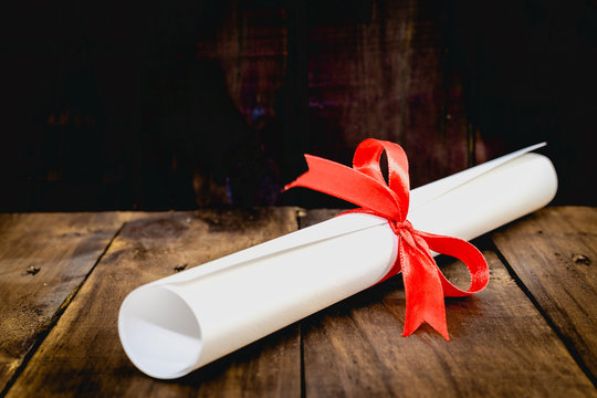 Mortarboard and graduation scroll,tied are a certificate with red ribbon, on wood background.