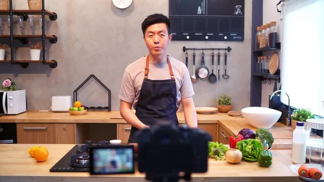 Young asian man in kitchen recording video on camera. Smiling asian man working on food blogger concept with fruits and vegetables in kitchen.