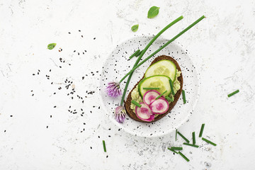 Toast avocado puree, cucumber, onion, onion flowers, radish and daikon on slices of dark bread on a light background with sesame seeds. Top view, copy space. - obrazy, fototapety, plakaty