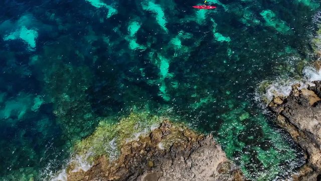 Overhead shot, flying from over a rocky shore above water. Two kayaking people appearing, beautiful blue and light blue pattern sea below them.