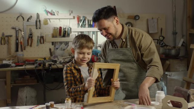 Medium shot of happy bearded father in apron teaching little son how to sand wooden picture frame