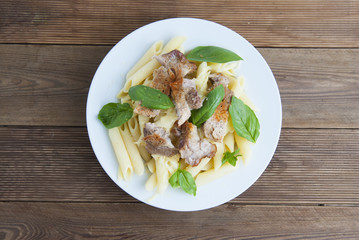 Cooked penne pasta isolated in white round plate, with basil leaves and pork meat, over wooden table, decorated with basil leaves. Top view.