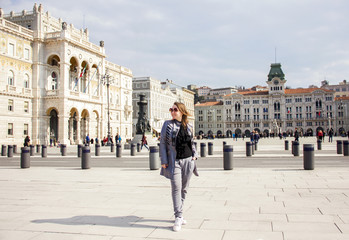 Obraz premium young attractive happy sunny girl in gray pants, cloak and sunglasses is standing fashion with gold backpack and posing on Trieste square background in Italy and smiling