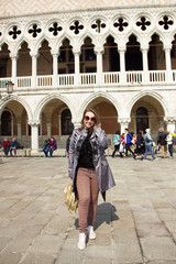 young attractive happy sunny girl in pants, gray cloak and sunglasses is standing fashion with gold backpack and posing on Venice square near the palace of the doges background in Italy and smiling