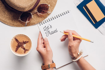 partial view of woman with notebook with lets travel lettering at tabletop with cup of coffee,...