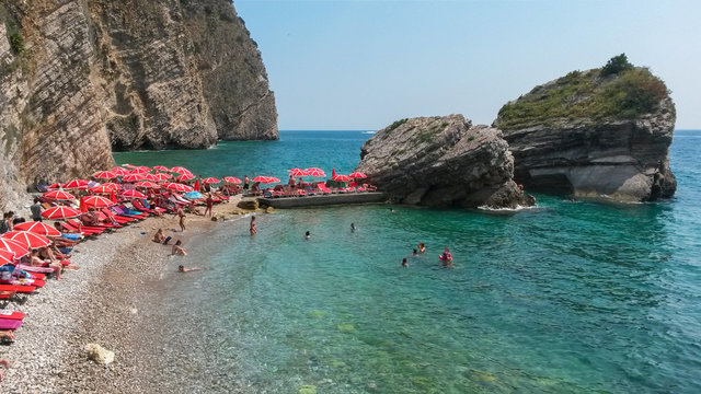 Panoramic view of pebble beach with clear azure blue water, layered rocks and red beach umbrellas, beautiful mediterranean Adriatic Sea coast, Montenegro