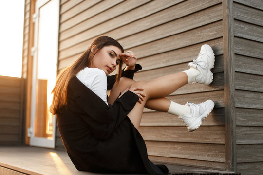 Beautiful young fashionable model woman in black stylish coat with sneakers sitting near a wooden wall at sunset
