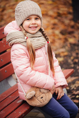 autumn portrait of smiling child girl sitting on bench in park in warm knitted hat and scarf, enjoying outdoor walk in sunny day