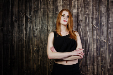 Fototapeta premium Portrait of a beautiful redheaded girl in black top and black skirt posing in the studio next to the wooden wall.