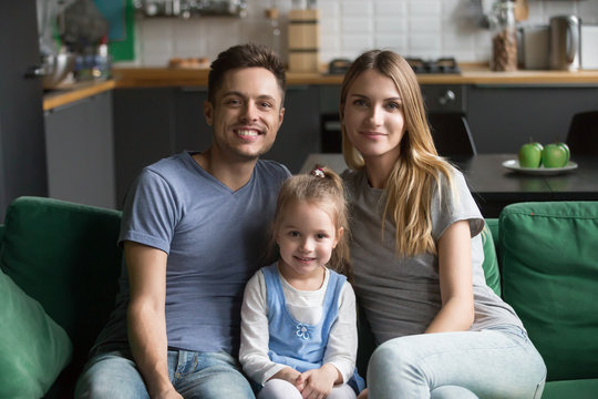 Portrait of happy healthy loving family of three concept, mom and dad posing with kid preschool daughter bonding together, young smiling parents sitting on sofa with child girl looking at camera