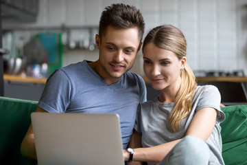Millennial happy couple shopping together, reading internet news or relaxing on sofa with laptop, young man and woman watching interesting video or studying online using computer at home on weekend