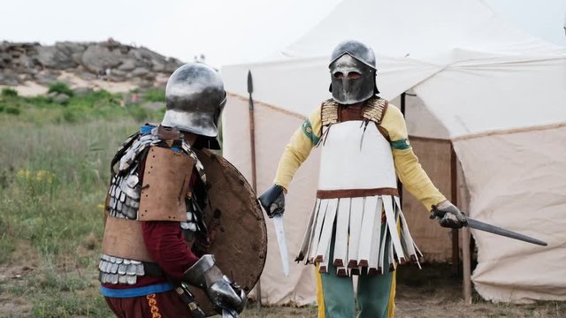 Two men in ethnic armor fight each other with swords and spear in a field.