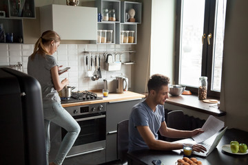 Young couple using laptop and smartphone in the kitchen, smiling man working online at home while...