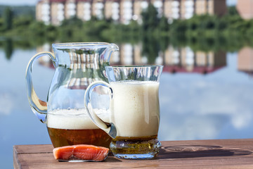 Jug with mug of beer and piece of fish on the table by lake