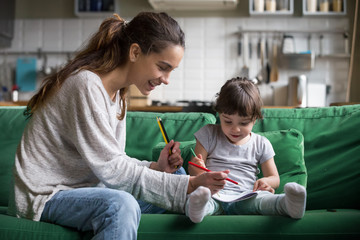 Smiling baby sitter and preschool kid girl drawing with colored pencils sitting on sofa together, single mother and child daughter playing having fun, creative family activities at home concept - obrazy, fototapety, plakaty
