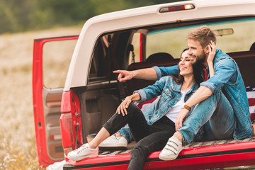 beautiful young couple relaxing in car trunk during trip and pointing somewhere