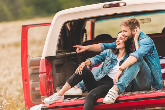 beautiful young couple relaxing in car trunk during trip and pointing somewhere - Powered by Adobe