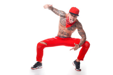 guy with tattoos in red jeans and a cap with a red kerchief around his neck and black hairstyles on a white background crouched and shows tongue