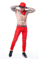 guy with tattoos in red jeans and a cap with a red kerchief around his neck and black hairstyles on a white background holds his hands behind his head