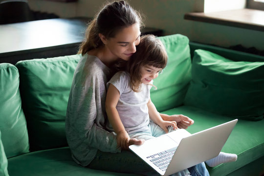 Happy Mother With Little Kid Daughter Having Fun Online With Application, Watching Video Cartoons Or Playing Game On Laptop Together, Young Babysitter Or Nanny Teaching Child Girl To Use Computer