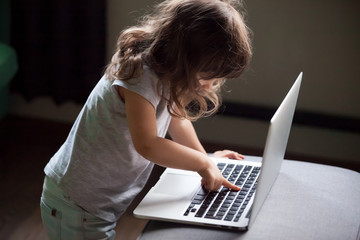 Curious smart little girl typing on laptop alone, clever cute child using computer online without...