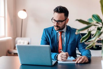 Handsome businessman in eyeglasses is making notes while working on a laptop